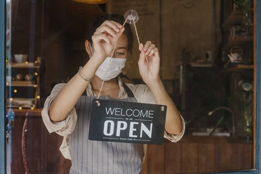 Young barista flips “Open” sign on café door while wearing mask